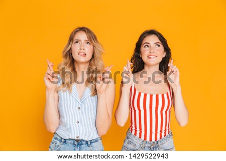 Portrait of two concentrated blonde and brunette women 20s in summer wear dreaming while keeping fingers crossed isolated over yellow background