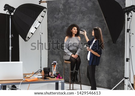 Portrait of female makeup artist applying cosmetics to african american woman during photo shooting with professional camera and softbox in studio