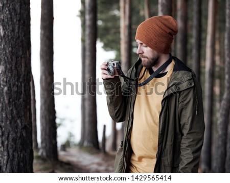Young man in hat and jacket looking at camera in hands and taking photo in gloomy slender forest