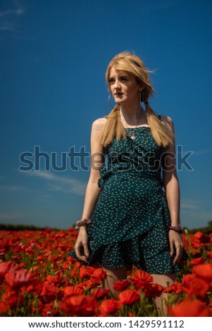 A blonde haired young woman wearing a spotty/polka dot dress walking through a poppy field, facing to the side