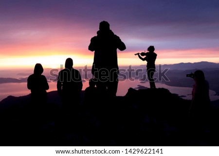 Multi-colored sunset. Silhouette of a woman with a violin. Silhouettes of people on top of a mountain overlooking the sea.
