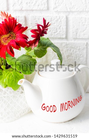 White tea pot with the signature Good morning, on the background of red gerbera flowers in a flowerpot of lace. Background white brick wall. Cozy morning, hot tea, breakfast
