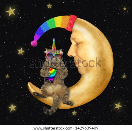 The cat unicorn in sunglasses and headphones is listening to music from a smartphone on the comfortable moon in a knitted hat. Stars background.