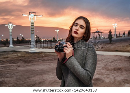 young beautiful girl in Burgundy stockings and gray coat stands with vintage camera at sunset