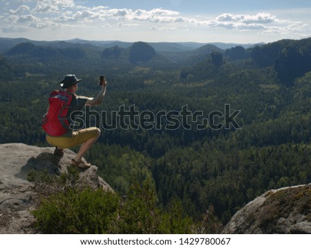 Hiking man with backpack, hat and hand camera sit on the mountain cliff. Phone  photographer taking picture of forest, sky and clouds on his camping trip. Sportsman travel alone