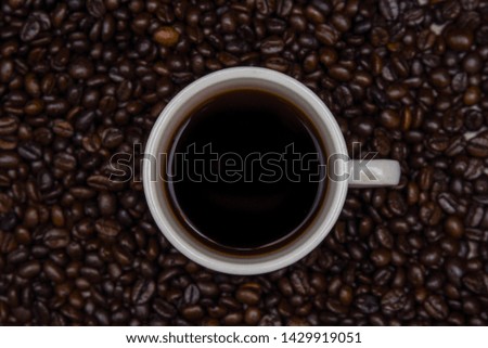 White coffee mug with black coffee water in a glass, placed on a table filled with coffee beans with a dark brown background.