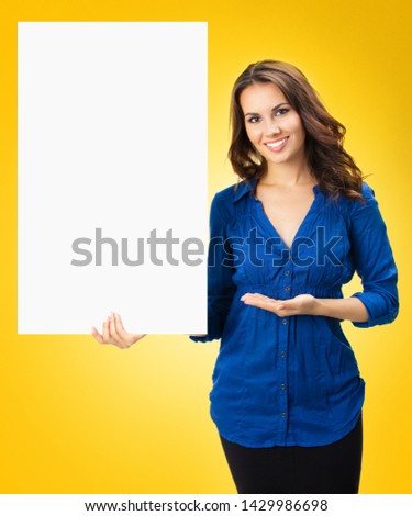 Happy smiling young woman showing blank sign board with empty copy space, isolated over yellow orange  background. Business and ad concept. Brunette model in blue confident clothing at studio picture.