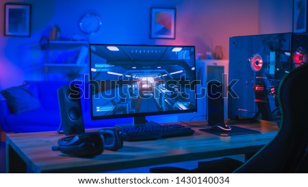 Powerful Personal Computer Gamer Rig with First-Person Shooter Game on Screen. Monitor Stands on the Table at Home. Cozy Room with Modern Design is Lit with Blue and Neon Light.