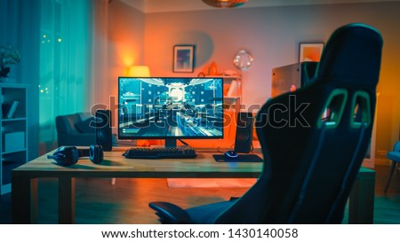 Powerful Personal Computer Gamer Rig with First-Person Shooter Game on Screen. Monitor Stands on the Table at Home. Cozy Room with Modern Design is Lit with Warm and Neon Light.