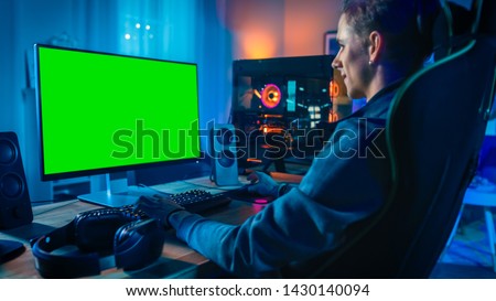 Excited Gamer Playing Online Video Game with a Mock Up Green Screen on His Powerful Personal Computer. Room and PC have Colorful Neon Led Lights. Cozy Evening at Home.