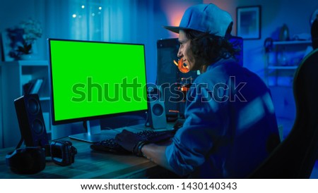 Professional Gamer Playing Online Video Game on His Powerful Personal Computer with Colorful Neon Led Lights. Green Screen Mock Up. Young Man is Wearing a Cap. Living Room with Warm Lamps. Evening.