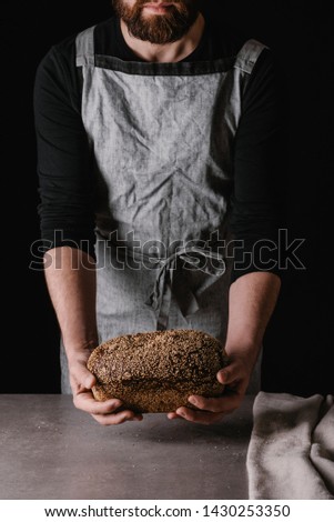 A man in an apron puts a loaf of fresh rye bread on the table. Black background.