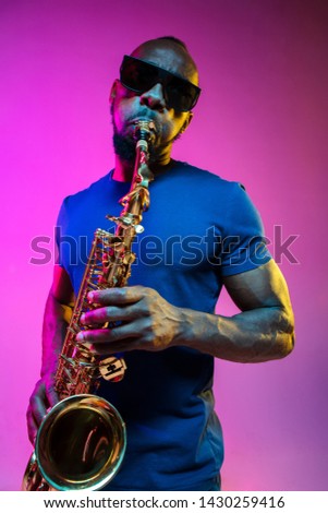 Young african-american jazz musician playing the saxophone on pink studio background in trendy neon light. Concept of music, hobby. Joyful attractive guy improvising. Colorful portrait of artist.