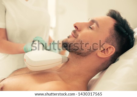 Calm young man lying with his eyes closed and cosmetologist in rubber gloves removing hair with laser tool Royalty-Free Stock Photo #1430317565