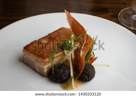 Roasted pork belly with deep fried wild mushroom balls, served with a pancetta crisp. Presented on a white plate on a dark wood table.
