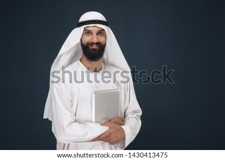 Half-length portrait of arabian saudi businessman on dark blue studio background. Young male model using tablet or gadget. Concept of business, finance, facial expression, human emotions, technologies