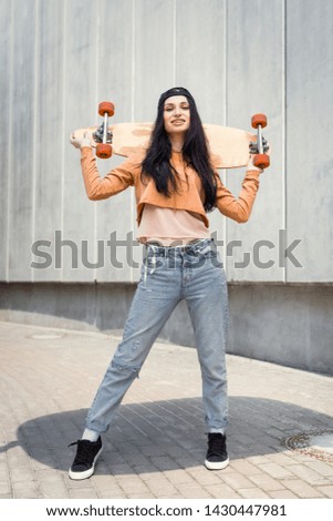 cheerful woman in casual wear standing near concentrate wall, holding skateboard behind back