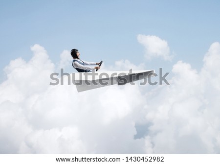 Aviator having fun in aircraft. Pilot in leather helmet and goggles driving paper plane in cloudy blue sky. Funny man flying in small paper airplane. Cloudscape background with fluffy clouds.