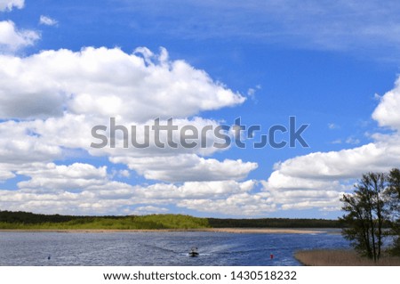 A view of a vast lake or river with many waves reflecting the white puffy summer clouds with some trees, reeds, and a forested coast with a boat swimming quickly across the surface of the reservoir