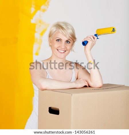 Portrait of a young casual woman with cardboard box and paint roller in front of half yellow painted wall