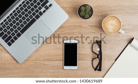 On the workplace, a laptop and a smartphone, an open notebook and glasses, a cup of coffee and a pot of cactus. View from above.