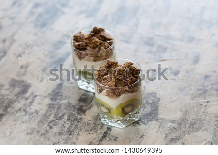 Granola, kiwi, banana and Greek yogurt in two glass cups on gray concrete, copy space. Fitness, figure, body and healthy food