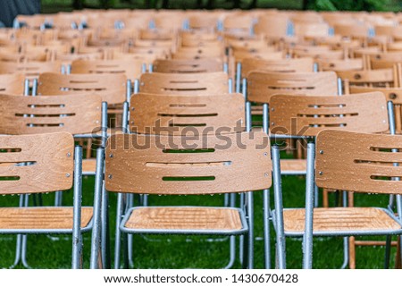 Lot of wooden chairs stand outside in the park in the rain. Empty auditorium, green grass, trees and chairs with water drops.