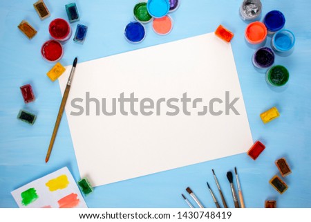 Watercolor, acrylic, brushes and a white sheet of paper on a blue background. Flat lay, top view. Minimalistic desktop of the artist. Preparation for drawing.