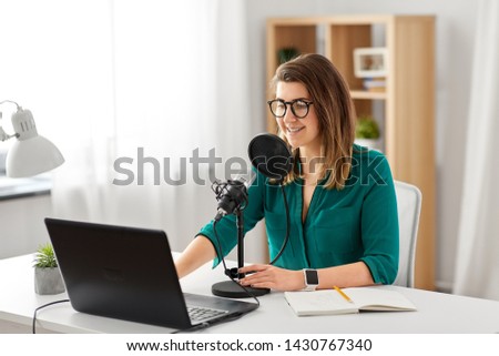technology, mass media and people concept - woman in glasses with microphone and laptop computer talking and recording podcast at studio