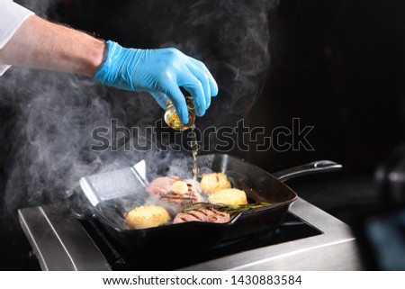 The chef's hand in glove over a frying pan with filet mignon and grilled vegetables is pouring white wine over the dish during cooking on the stove.