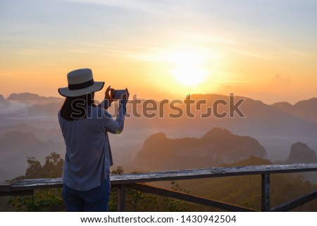 backside young asian woman feeling happy wearing hat with photography by smart phone mobile stand resting on tropical mountain at Doi Ta Pang (khao thalu) travel in chumphon thailand