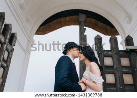 Beautiful newlyweds stand on the background of an ancient stone arch with wooden doors. Stylish bride in a hat and a young bride with curly hair holding hands near the palace.