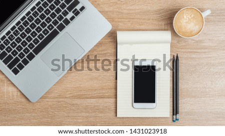 
A laptop, a smartphone on an open notepad, a couple of pencils and a cup of coffee on the desktop. View from above.