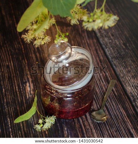 A jar of wild strawberry jam with linden flowers scattered across the frame on a dark wooden background