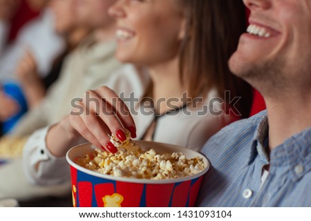 Selective focus of female hand taking delicious cheese popcorn in cinema. Cheerful pair watching new interesting comedy and eating tasty snack. Concept of relaxation and entertainment.