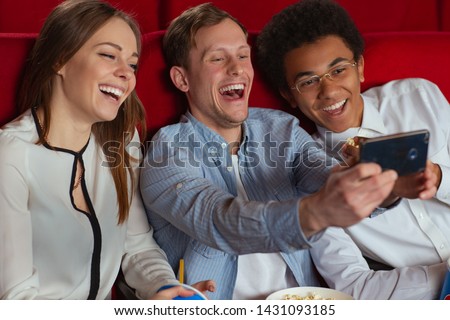 View from side of three friends sitting in cinema and taking selfie together. Cheerful company looking at camera and laughing before watching film. Concept of entertainment and fun.