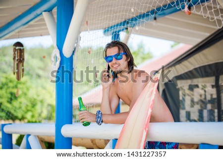 Young attractive smiling man surfer talking on the smartphone and holding beer in the other hand. Caravan camping. Summer vacation recreation photo.