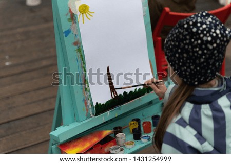 Kid leisure concept. Child in a black sparkled hat and a striped jacket is painting with gouache on open-air lesson close up, back view. Art school or workshop class.