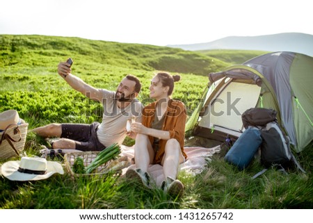 Young and cheerful couple making selfie photo during the picnic at the campsite, traveling high in the mountains during the sunset
