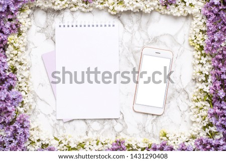 Mobile phone mock up, notebook and frame of white and lilac flowers on marble table in flat lay style.