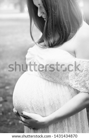 Pregnancy, love, people and expectation concept - happy pregnant woman posing over green natural background in white dress, holding belly with both hands. Black and white photo.