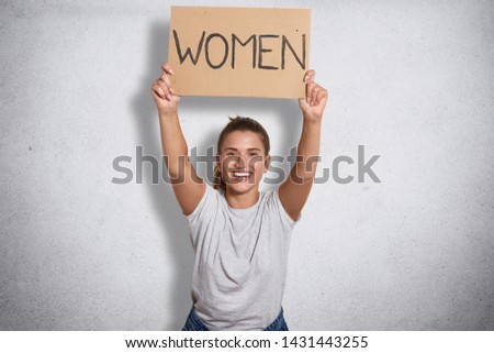 Cheerful good looking feminist smiling sincerely, raising inscription women in both hands, ready to public activity, peaceful demonstration, being in high spirits, wearing casual white clothes.