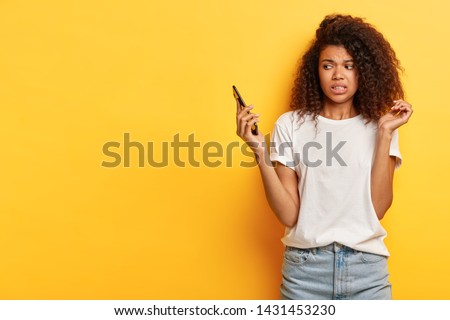 Beautiful female with frizzy Afro hairstyle, looks in displeasure at cell phone, receives call from former boyfriend, holds mobile phone on distance, ignores communication, afraids of something Royalty-Free Stock Photo #1431453230