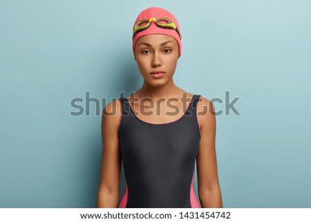 Photo of serious female holiday maker prepares for swimming marathon on beach, looks confidently, dressed in swimsuit, goggles on head, leads healthy lifestyle, has slim figure, likes active rest
