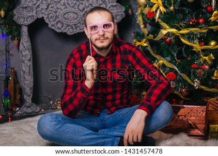 The guy in the red shirt is holding glasses on a stick. A bearded man sits on a white carpet on the background of a Christmas tree. Christmas decor