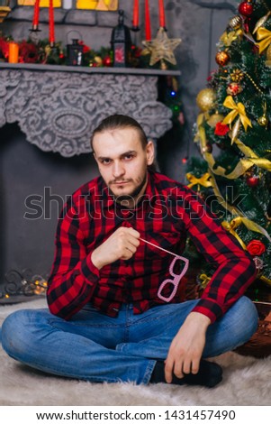 The guy in the red shirt is holding glasses on a stick. A bearded man sits on a white carpet on the background of a Christmas tree. Christmas decor