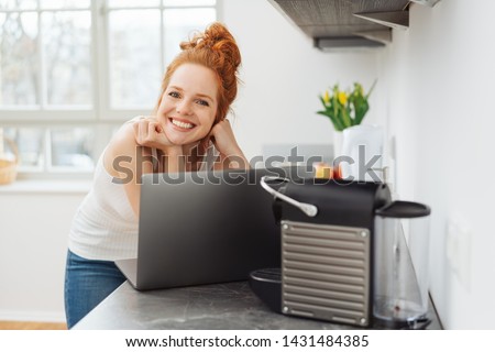 Young woman relaxing in a kitchen with a laptop computer balanced on the counter looking over the top to smile happily at camera