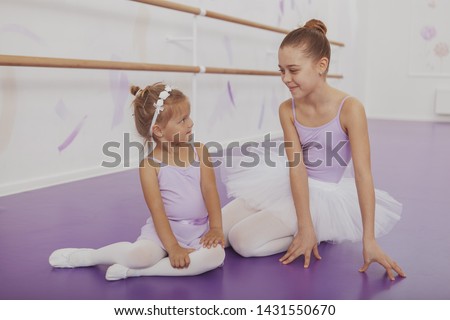 Two lovely young ballerinas smiling at each other, resting on the floor after dancing at ballet school. Adorable little ballerina and her sister wearing leotards and skirts relaxing after class