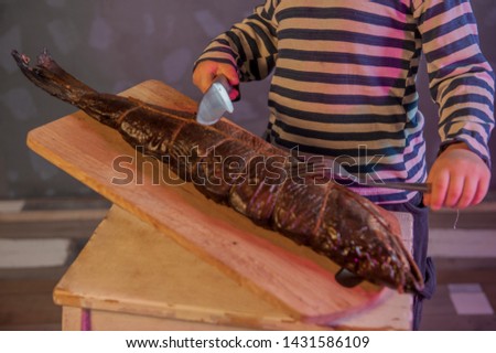 Little blond boy in cook's hat shows big smoked fish. Child tries fish with knife and fork. Kid wants to cut and cook fish. Concept of organic food.