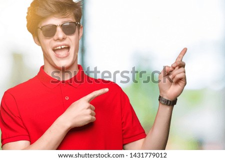 Young handsome man wearing sunglasses over isolated background with a big smile on face, pointing with hand and finger to the side looking at the camera.